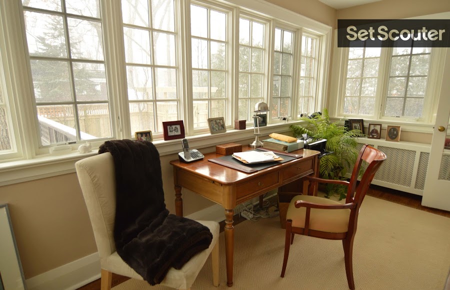 Large wall-to-wall picture windows allow natural light to illuminate Jackie's sunroom-turned-office space. Small details, like the corner plant and littered photo frames keep the space looking traditional and relaxed. This is the perfect room to showcase a character that’s both hard working and family-oriented.