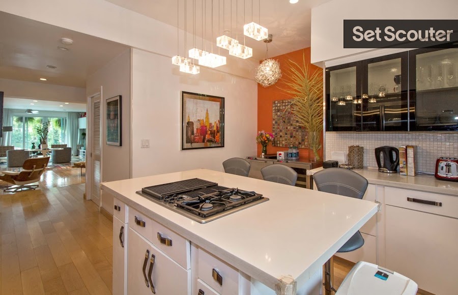 A quaint and trendy kitchen highlights an island with a counter top stove. Granite counters and stainless steel appliances are sleek and clean in this vibrant space. The addition of an orange accent wall, contemporary pendant lighting and glass cupboards have this kitchen beaming with charm and energy.