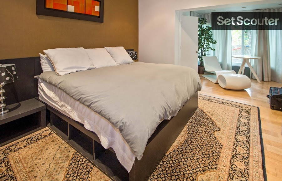 The master bedroom is quiet in decor with a light brown accent wall and large patterned carpet. Tan hardwood floors and large windows add warmth to the room while indoor plants and a modern seating area keep the space crisp and organic.