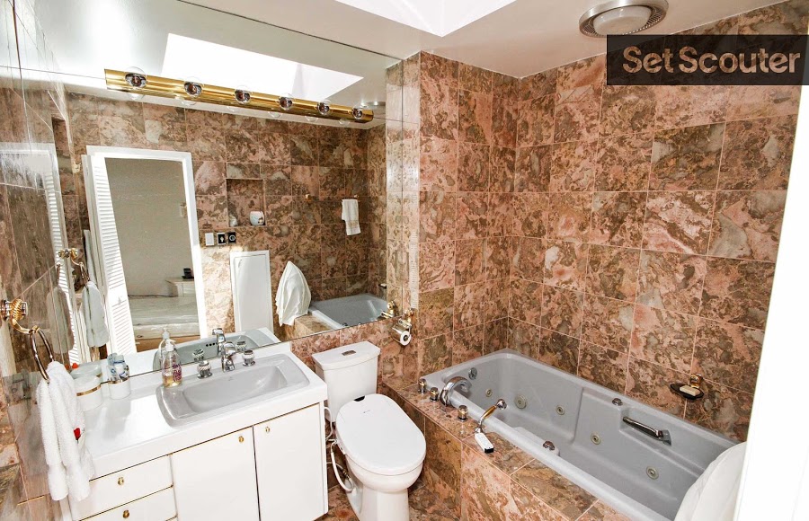 A marble-tile covered bathroom makes for a unique, at-home spa. With bright, white amenities, a jacuzzi and gold-coloured faucets, this space exudes wellness and luxury. A skylight lengthens the room while a large bathroom mirror opens it up.