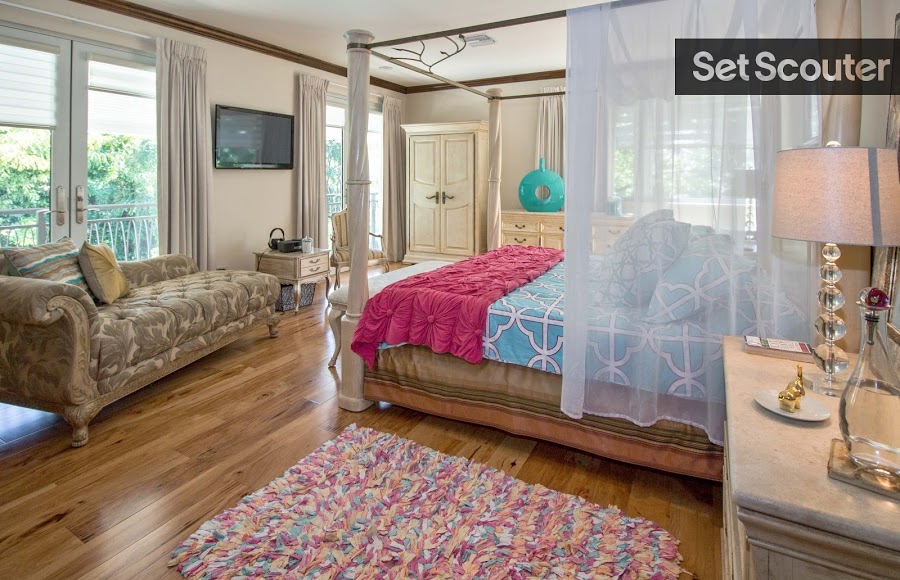 The upstairs bedroom has graceful charm and some great vintage pieces. Pops of colour throughout this neutral space contrast beautifully with rustic, traditional items like the chaise, wardrobe and side tables. A canopy bed is the center of attention while the room opens up to a balcony with classic french doors behind subtle white curtains.